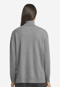 Frau mit langen, dunklen Haaren, die einen lockeren grauen Rollkragenpullover und schwarze Jeans trägt, von hinten auf einem weißen Hintergrund gezeigt.