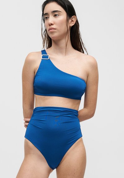 Γυναίκα με μακριά σκούρα μαλλιά που φοράει μπλε μπικίνι τοπ με έναν ώμο και αγκράφα και μπλε μπικίνι σλιπ με ψηλή μέση, στέκεται με τα χέρια πίσω από την πλάτη.