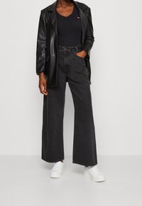 Svart läderblazer över en svart v-ringad topp, tillsammans med vida svarta jeans och vita sneakers. Klädseln har en figursydd silhuett och kontrasterande texturer.