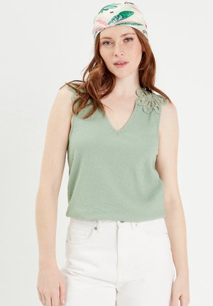 Débardeur vert menthe avec une finition texturée et un col en V, présentant un détail en crochet décoratif sur l'épaule gauche. Associé à un pantalon blanc.