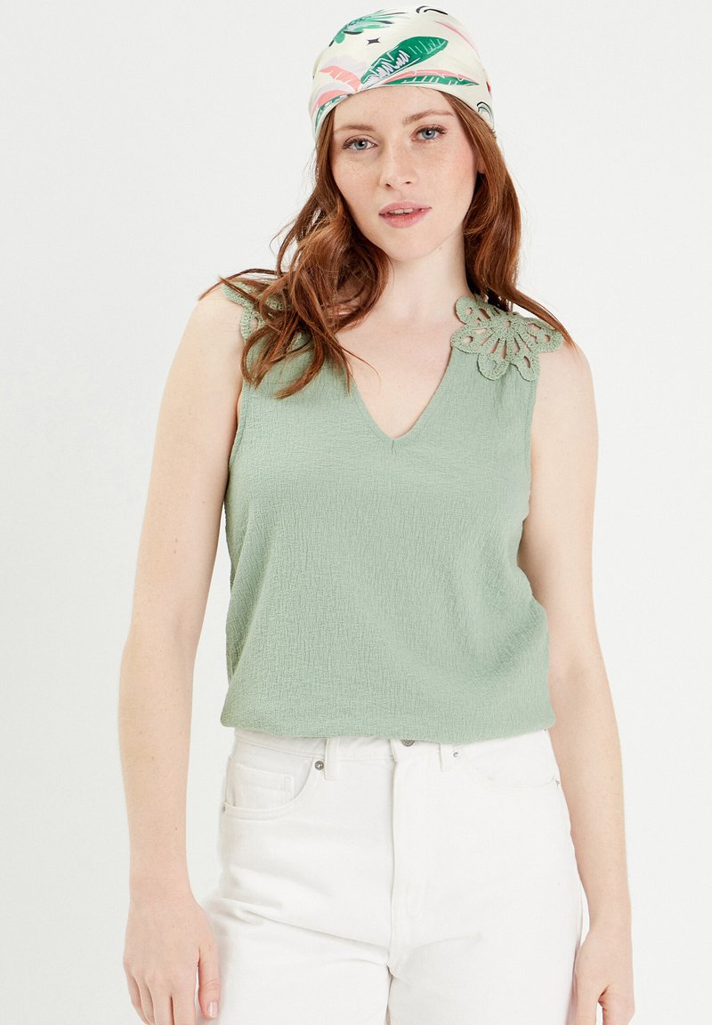 Débardeur vert menthe avec une finition texturée et un col en V, présentant un détail en crochet décoratif sur l'épaule gauche. Associé à un pantalon blanc.