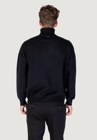 Zwarte gebreide pullover met een hoge kraag, geribbelde manchetten en verstelbare tailleband. Soepele textuur, relaxte pasvorm, minimale branding bij de hals.