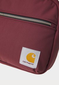 Bordeaux nylon bag med en teksturert overflate, med en sølvglinsende glidelås og et hvitt merke med en gul logo i nederste hjørne.
