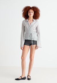 Haut enveloppant gris clair à manches longues en tissu texturé avec une taille à nouer, associé à un short sombre et des ballerines noires. Fond uni.