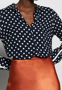 Mørkeblå skjorte med knapper og hvide prikker; har krave og lange ærmer. Kombination med en skinnende orange satin nederdel.