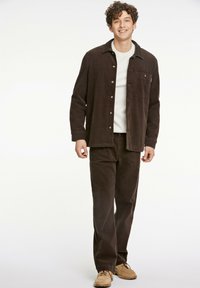 Brun manchester skjortjacka med knappfront, tillsammans med matchande bruna manchesterbyxor och beiga skor. Bakgrunden är enkel vit.