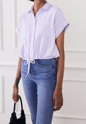 Henkilö, jolla on vaaleanvioletti lyhythihainen napillinen paita, jossa etutaskut ja solmittava vyötärö, yhdistettynä sinisiin farkkuihin, ja joka pitää mustaa käsilaukkua.