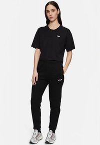 Zwarte cropped T-shirt en bijpassende joggers, beide met een wit FILA-logo. Model draagt metalen sneakers met accenten in grijs en wit.