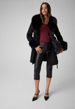 Femme portant un manteau noir à col en fourrure, un haut bordeaux, un pantalon court en cuir noir, des talons noirs et un grand sac à main noir.