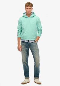 Felpa verde menta con tasca frontale, indossata sopra una camicia a righe blu chiaro, abbinata a jeans blu scoloriti e scarpe da ginnastica beige.