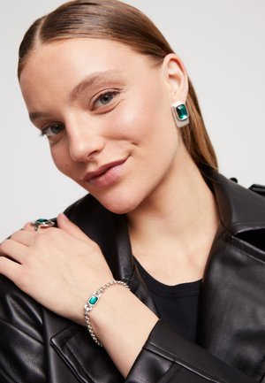 Joyería de plata con piedras preciosas verdes; un anillo y una pulsera en una mano, usados con una chaqueta de cuero negro y una camisa negra. Pelo corto castaño visible.