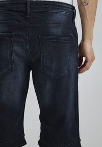 Shorts en denim foncé avec une taille ajustée, des coutures de poches visibles et une légère décoloration, présentant un ourlet retourné et deux poches arrière.