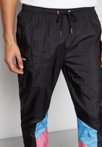 Man wearing black track pants with blue and pink color blocks at the calves and a drawstring waist, standing with hands relaxed.