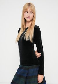 Bluză neagră din catifea cu mâneci lungi și decolteu rotund, combinată cu o fustă în carouri albastru și verde. Textură netedă și croială mulată.