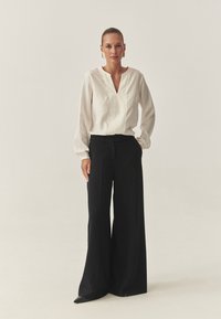 Weiße Bluse mit langen Ärmeln, V-Ausschnitt und Falten, kombiniert mit hochtaillierten, schwarzen, weit geschnittenen Hosen. Einfaches Design und glatte Textur.
