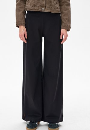 Osoba nosząca szerokie czarne spodnie, ciemne trampki i krótki brązowy płaszcz, z rękami swobodnie opuszczonymi wzdłuż ciała.