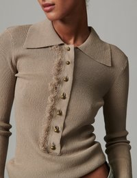 Γυναίκα που φοράει ένα μπεζ ριμπ πλεκτό τοπ με μυτερό γιακά και πατιλέτα με κουμπιά που διαθέτει έξι ορειχάλκινα κουμπιά και διακοσμητικό φθαρμένο τελείωμα.