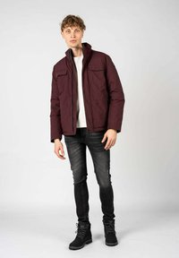 Giacca puffa bordeaux con due tasche sul petto, chiusura zip e bottoni a scatto. Indossata con jeans neri e stivaletti neri con lacci, design minimalista.