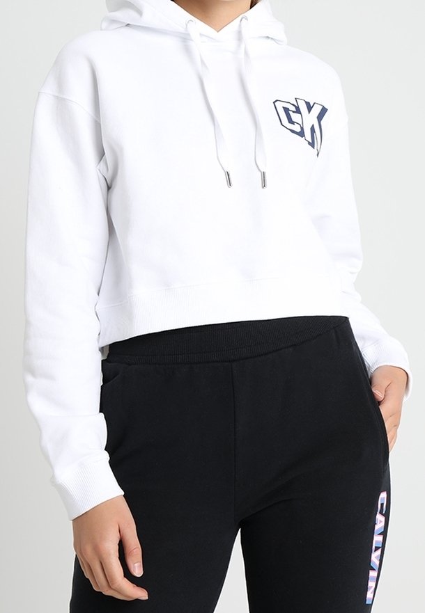 Felpa bianca corta con cappuccio, con il logo navy "CK" sul lato sinistro del petto. Abbinata a pantaloni sportivi neri con il testo "CALVIN".