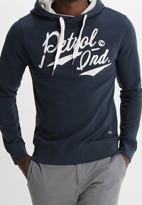 Hombre vistiendo una sudadera con capucha azul marino y cordones blancos, con el logo "Petrol Ond.", combinado con pantalones gris claro, con las manos en los bolsillos.