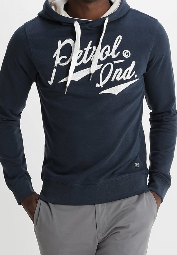 Hombre vistiendo una sudadera con capucha azul marino y cordones blancos, con el logo "Petrol Ond.", combinado con pantalones gris claro, con las manos en los bolsillos.