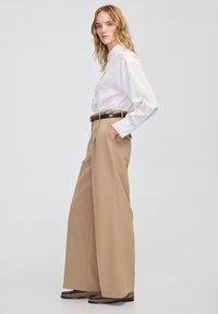 Wit gestreept shirt met lange mouwen, gecombineerd met hoog uitgerokken beige wijde broek en donkere patroon schoenen, tegen een effen achtergrond.