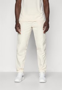 Pantaloni sportivi color crema con orli elastici, design affusolato e finitura liscia. Indossati con una maglia chiara abbinata e sneakers bianche.