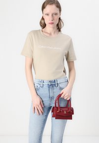 Camiseta de algodón beige con la inscripción "Calvin Klein Jeans" en letras blancas; jeans de mezclilla azul claro; mini bolso rojo con dos asas y acentos dorados.