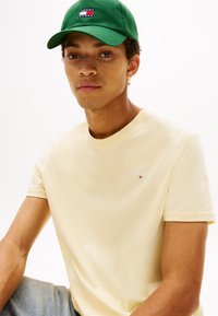 T-shirt en coton jaune à manches courtes, avec un petit logo blanc "TOMMY JEANS". Associé à une casquette verte affichant un logo multicolore.