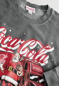 Pelēks džemperis ar vintage Coca-Cola dizainu, sarkaniem burtiem un priekšpusē redzamu Ziemassvētku vecīša ilustrāciju, kurš tur pudeli.