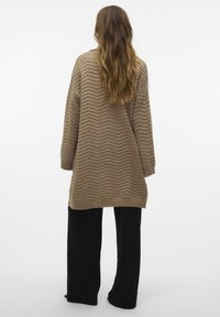 Übergroßer beiger Pullover mit einem Zickzackmuster, ausgestattet mit langen Ärmeln und einem lockeren Schnitt, kombiniert mit schwarzen weiten Hosen.