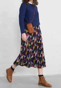 Maglione blu a maglia, gonna plissettata a fantasia con forme geometriche multicolori, stivali in pelle marrone e una piccola borsa a tracolla marrone.