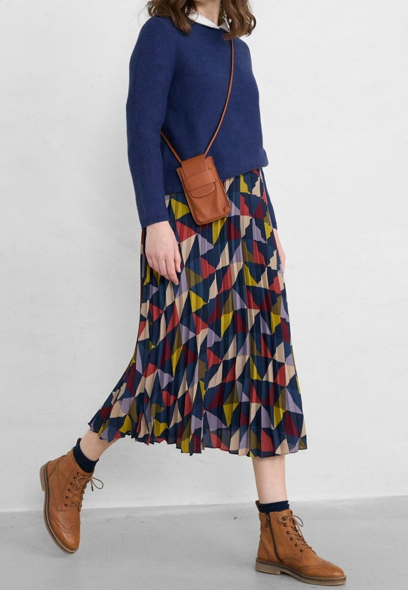 Maglione blu a maglia, gonna plissettata a fantasia con forme geometriche multicolori, stivali in pelle marrone e una piccola borsa a tracolla marrone.