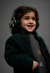 Child wearing black over-ear headphones, green fleece jacket, and gray sweatshirt with blue and gold embroidery. Smiling, with curly hair.