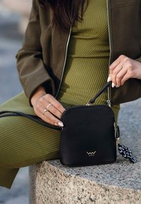 Černá kožená kabelka přes rameno s texturovaným povrchem, zlatým kováním a ozdobným střapcem. Kompaktní kulatý tvar se zipovým uzávěrem.