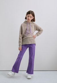 Sudadera beige con capucha y gráfico de calavera púrpura, combinada con jeans de campana púrpuras. La modelo lleva zapatillas blancas y se encuentra de pie contra un fondo neutro.