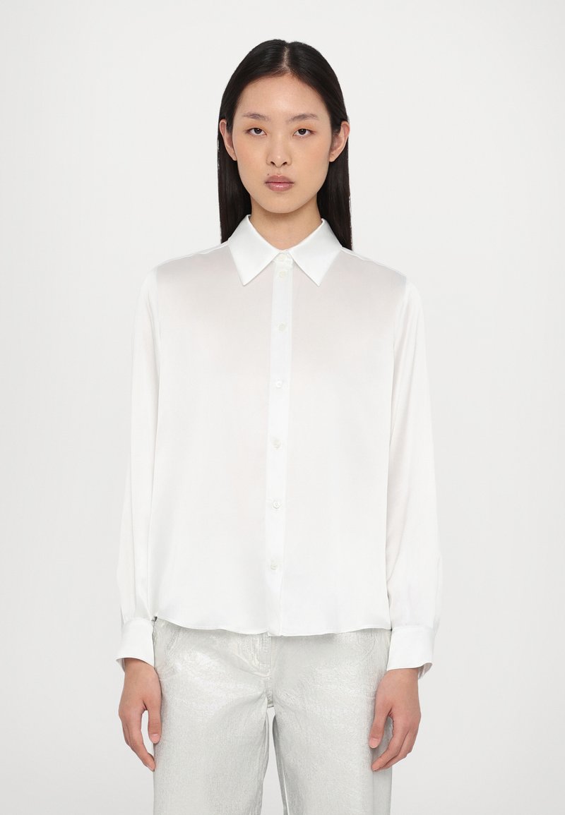 Witte lange mouwen blouse met een klassieke kraag, knoopsluiting aan de voorkant en een gladde textuur. Gecombineerd met glanzende zilveren broeken voor contrast.