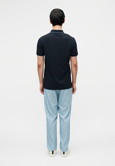 Μπλε-ναυτικό πόλο πουκάμισο με γιακά, συνδυασμένο με ανοιχτό μπλε παντελόνι με λεπτές ρίγες και λευκά αθλητικά παπούτσια, βλέποντας από πίσω σε απλό φόντο.