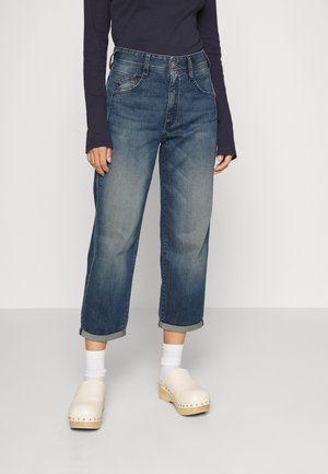 Persoon met opgerolde blauwe spijkerbroek, witte sokken, beige klompen en een donker topje met lange mouwen, staand tegen een effen achtergrond.