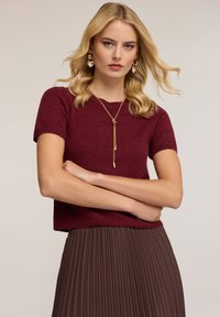 Bordový pletený top s krátkým rukávem a uvolněným střihem, v kombinaci s hnědými skládacími sukněmi. Má kulatý výstřih a hladkou texturu.
