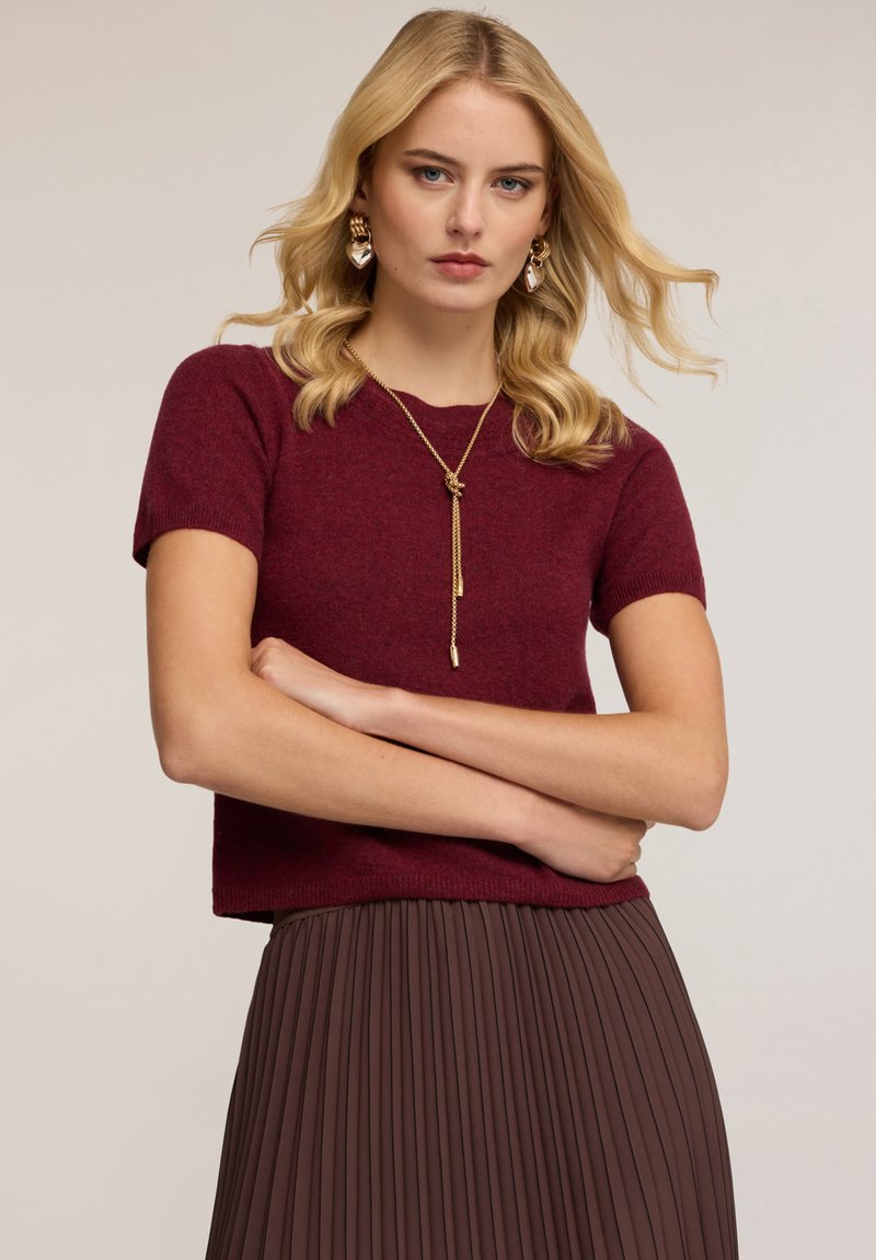 Bordový pletený top s krátkým rukávem a uvolněným střihem, v kombinaci s hnědými skládacími sukněmi. Má kulatý výstřih a hladkou texturu.