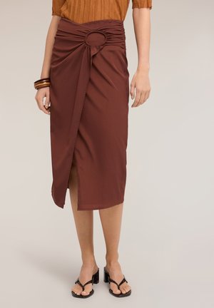 Vrouw draagt een bruine midirok met een geplooid rond detail in de taille, gecombineerd met zwarte sandalen met hakken en houten armbanden.