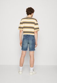Camiseta de manga corta a rayas en amarillo, azul marino y blanco, combinada con pantalones cortos de denim con dobladillo enrollado. Zapatillas blancas con detalles en tonos claros.