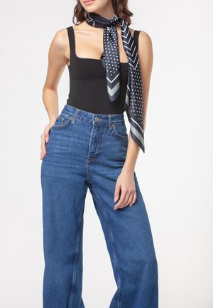 Donna che indossa jeans blu a vita alta e gamba larga, top nero senza maniche con scollo quadrato e una sciarpa di seta nera con motivo annodata intorno al collo.