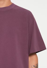 Lyhythihainen t-paita syvän purppuran värisessä puuvillassa, jossa on miehustakuvio ja sileä rakenne. Sisältää hienovaraisia ompeleita reunoilla.