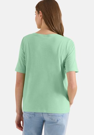 Donna con lunghi capelli castani che indossa una maglietta verde chiaro a maniche corte e jeans azzurri, vista da dietro.