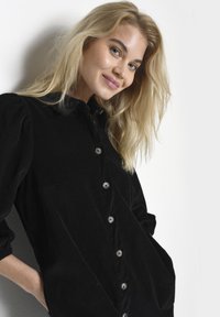 Abito camicia in corduroy nero con scollatura a collare, chiusura con bottoni frontale e maniche lunghe a sbuffo; presenta tasche laterali e un tessuto testurizzato.