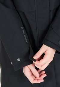 Svart vattentät jacka med en texturerad yta. Inkluderar justerbara ärmslut med tryckknappar och en etikett som säger "WPRO 10000" på ärmen.