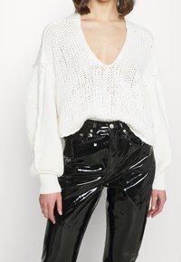 Weißer Strickpullover mit tiefem V-Ausschnitt und lockeren Ärmeln, kombiniert mit glänzenden schwarzen, hochgeschnittenen Hosen, die einen strukturierten Kontrast schaffen.