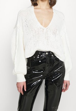Maglione bianco lavorato a maglia con scollatura a V profonda e maniche ampie, abbinato a pantaloni neri lucidi a vita alta, creando un contrasto di texture.